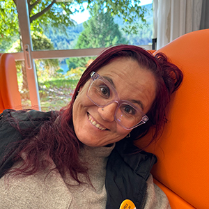 Eva, mujer adulta con cabello largo y rojizo, gafas y sonrisa amplia, sentada en una silla naranja