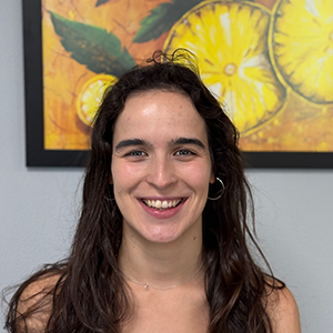 Maite, mujer joven con pelo largo oscuro y sonrisa alegre, con un cuadro brillante detrás